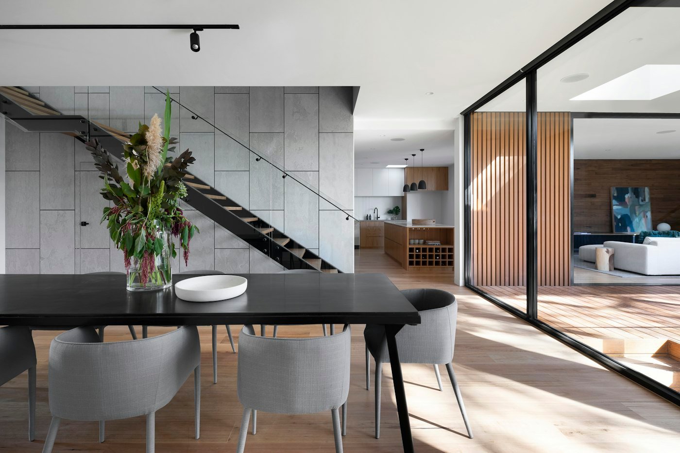 A black dining table with grey chairs, a stair with a glass rail, and a kitchen beyond